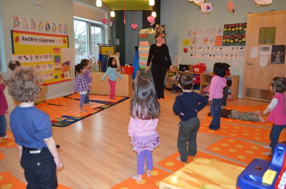 Children standing in a colorful classroom, engaged in a group activity led by an adult.